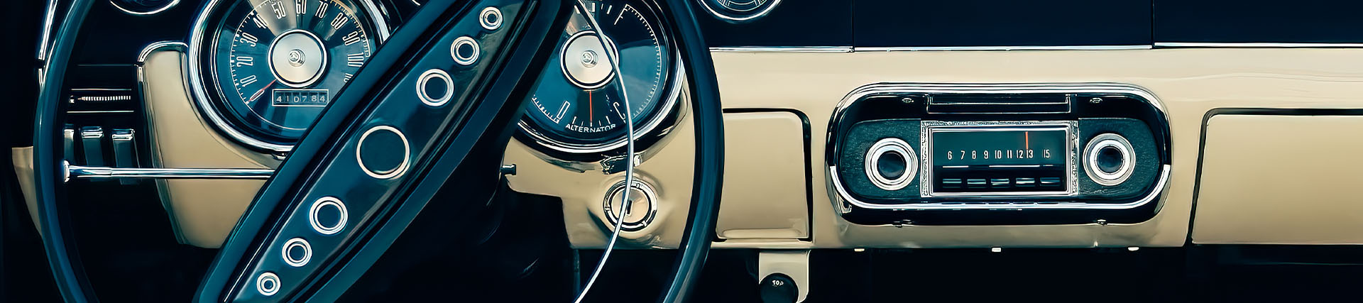 A short banner crop of a classic car dashboard with chrome gauges, a large steering wheel, and a vintage radio.