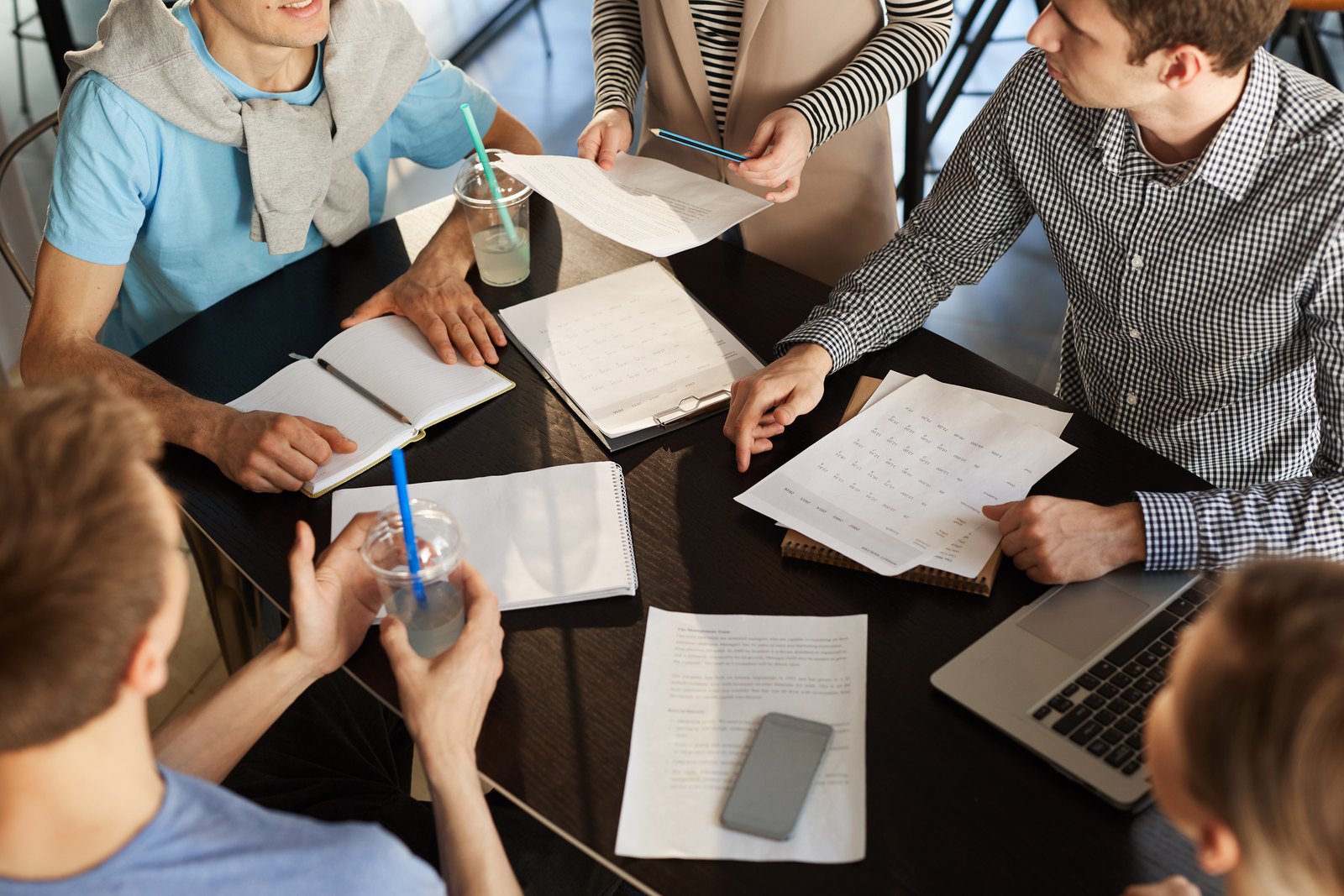 Seen from above, five people sit around a dark table covered with paper forms, notebooks and printouts, passing pages between them.