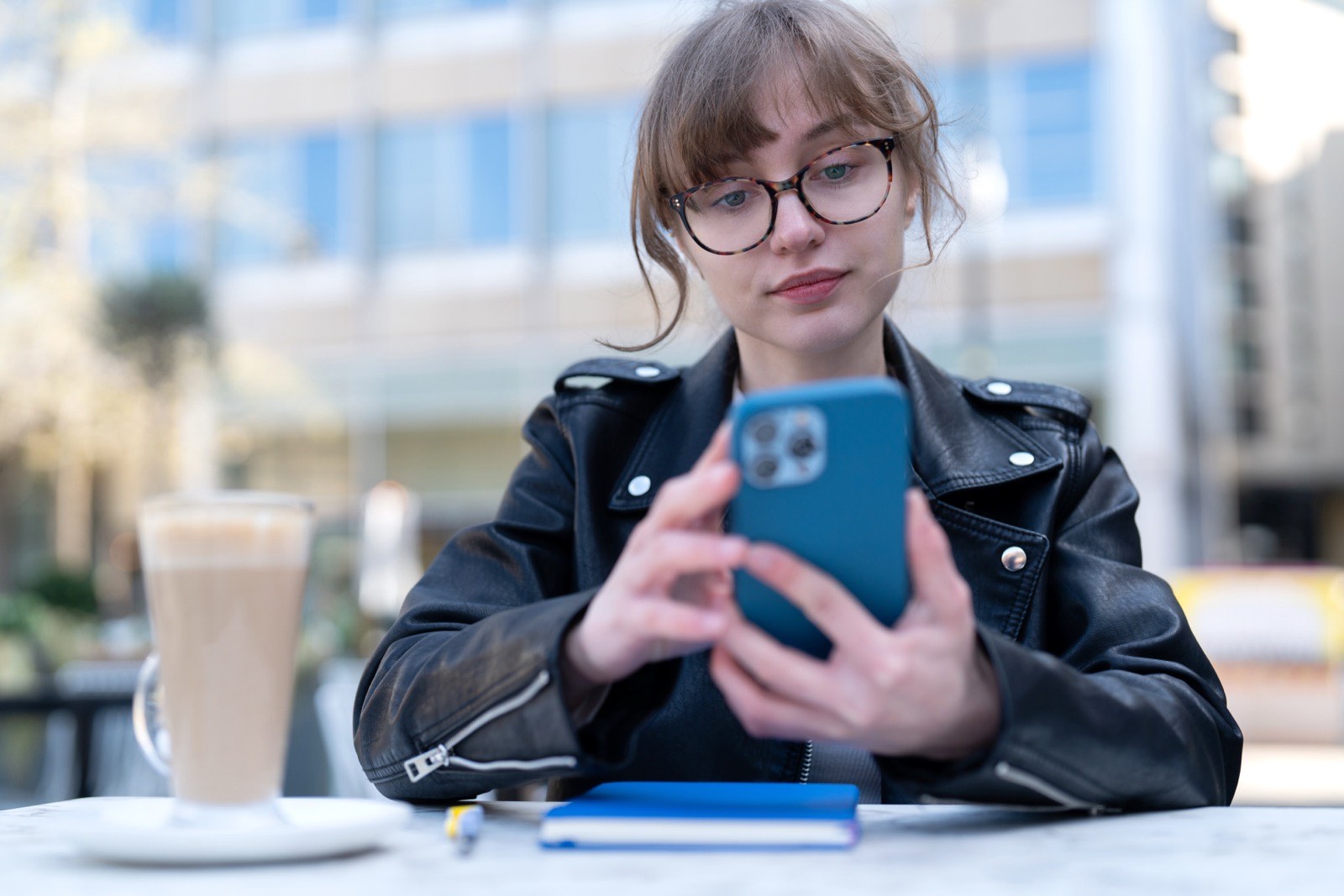 Young woman sitting at an outdoor cafe table, reading her phone.