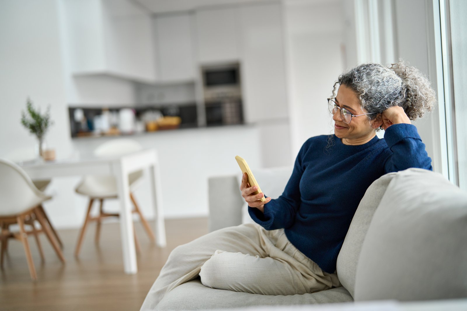 A woman with grey curly hair and glasses sits back on a sofa at home, reading something on her phone.