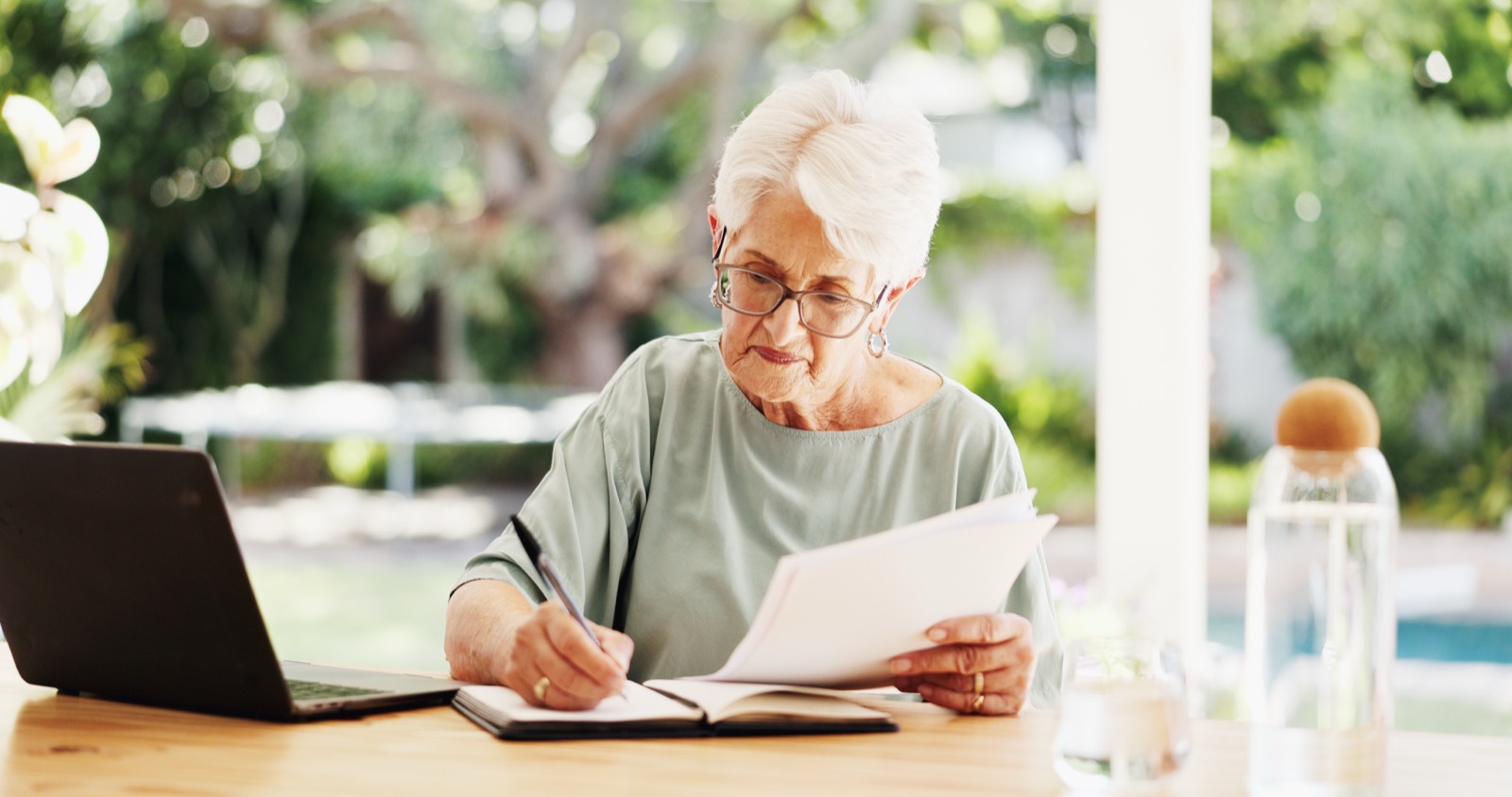 An older club volunteer writing notes by hand in a notebook beside a laptop and a stack of papers.