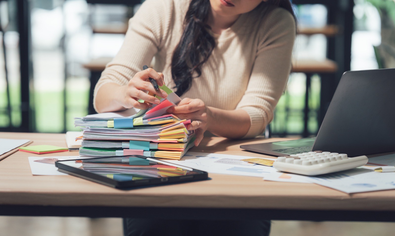 Woman at table with laptop, and a stack of paper documents.