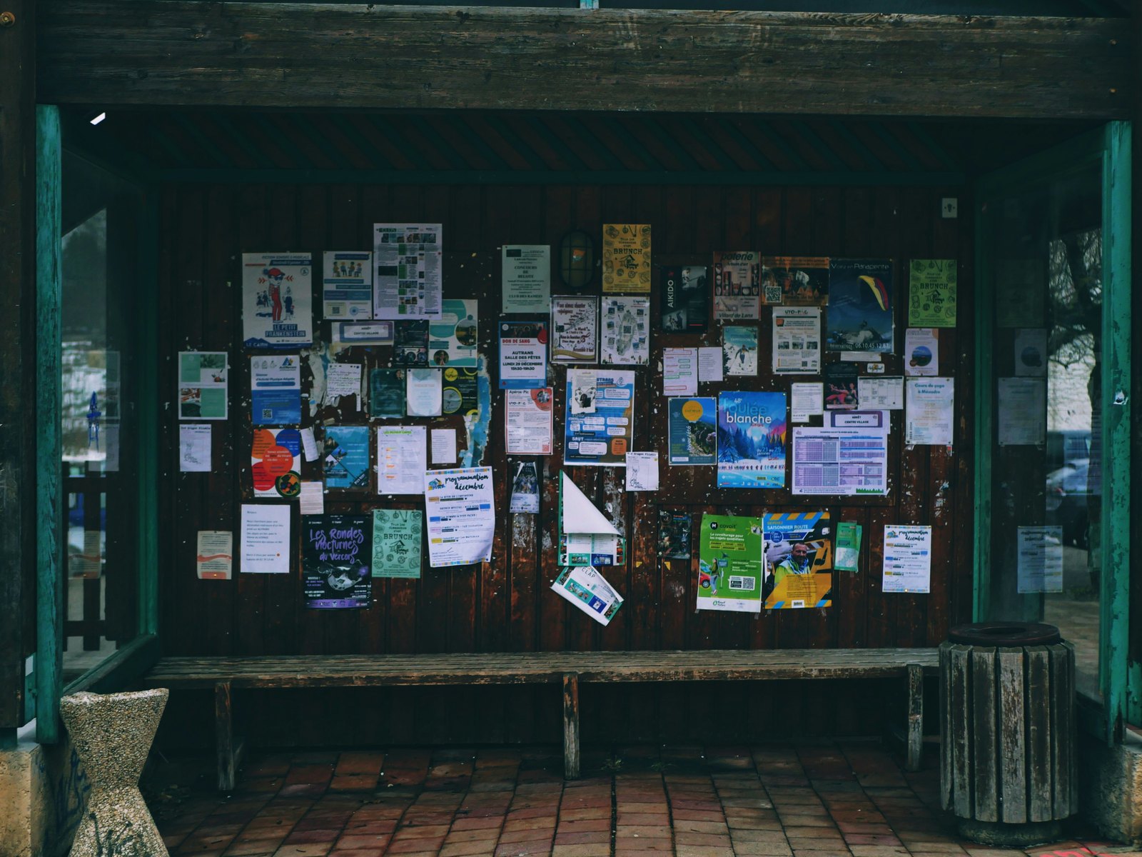A community noticeboard in a wooden shelter, covered in posters and flyers, with a bench beneath it.